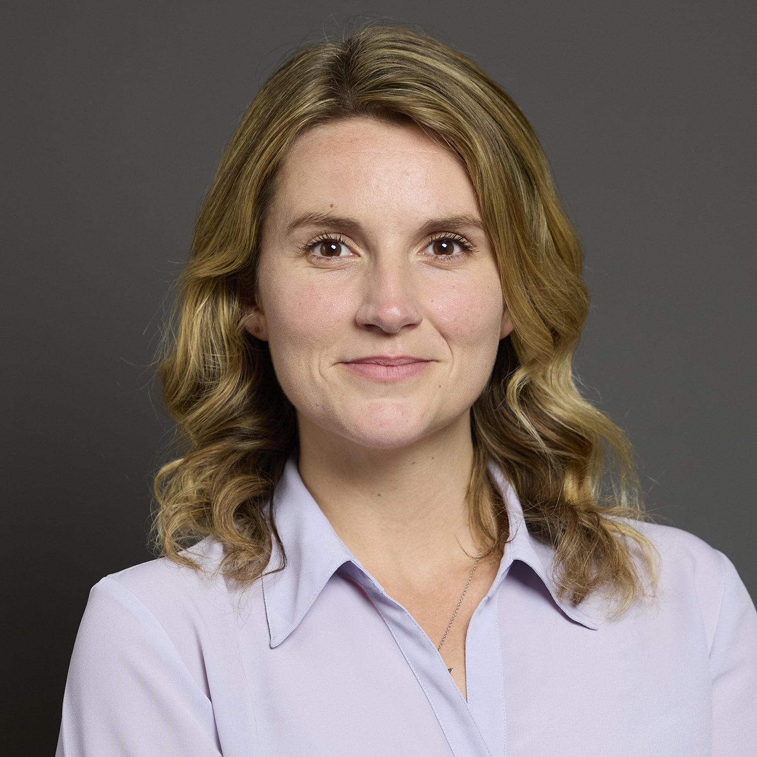 Portrait of a woman with wavy blonde hair wearing a light blue collared shirt against a gray background.
