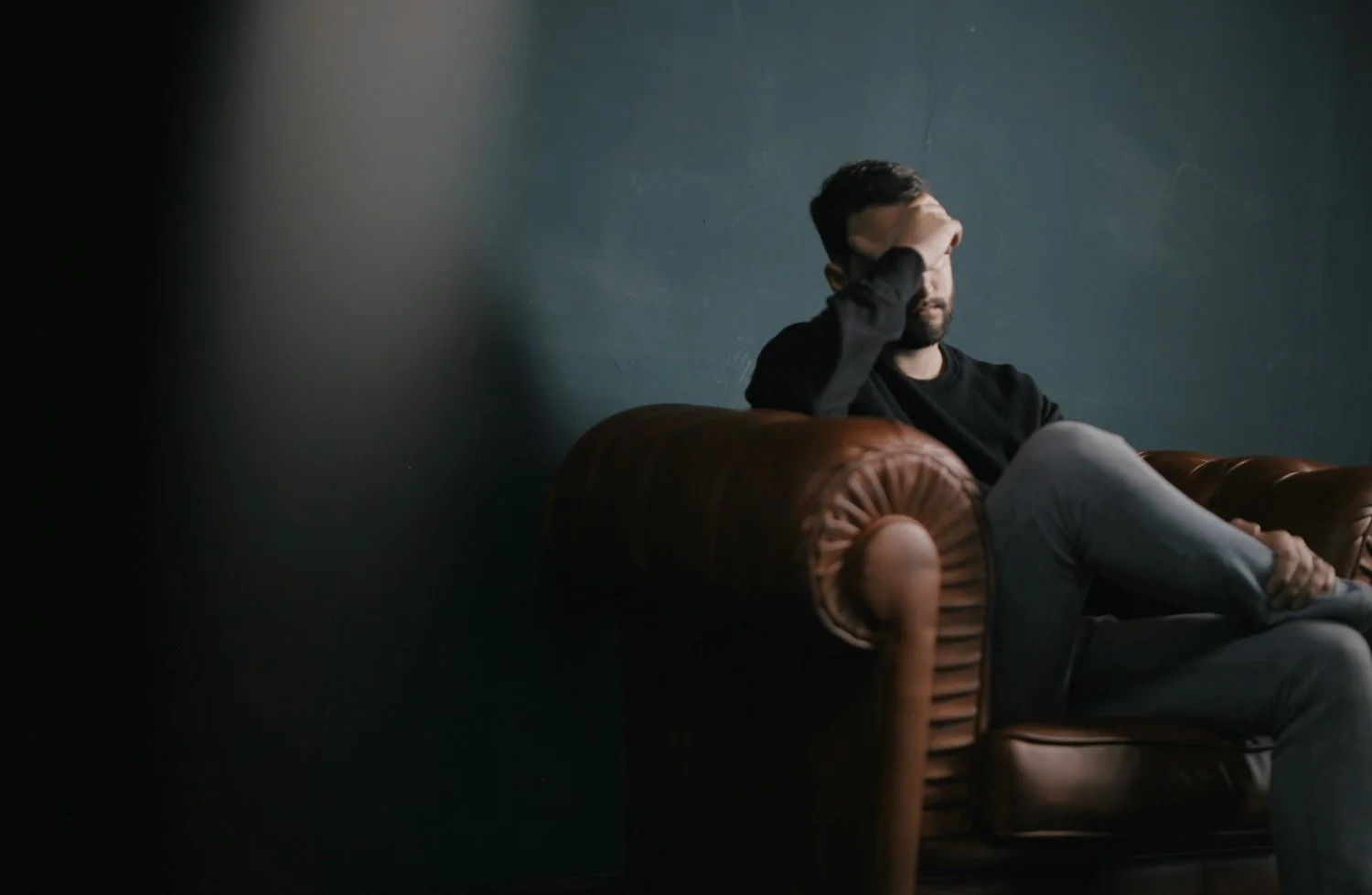 Man sitting on a brown leather couch with hand covering face